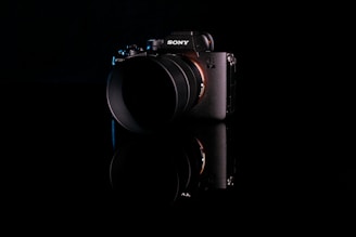 A high-end digital camera with a lens, featuring the Sony logo on a dark, reflective surface. The camera body is primarily black with a subtle sheen, highlighting its sleek design and modern technology.