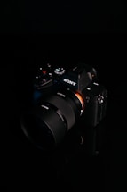 A digital camera with a sleek black design is placed against a dark background. The brand logo is clearly visible on the body of the camera, and various control buttons and dials are present. The lens is prominent, featuring detailed markings.