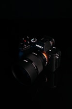 A digital camera with a sleek black design is placed against a dark background. The brand logo is clearly visible on the body of the camera, and various control buttons and dials are present. The lens is prominent, featuring detailed markings.