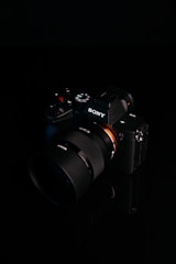 A digital camera with a sleek black design is placed against a dark background. The brand logo is clearly visible on the body of the camera, and various control buttons and dials are present. The lens is prominent, featuring detailed markings.