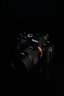 A digital camera with a sleek black design is placed against a dark background. The brand logo is clearly visible on the body of the camera, and various control buttons and dials are present. The lens is prominent, featuring detailed markings.