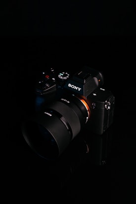 A digital camera with a sleek black design is placed against a dark background. The brand logo is clearly visible on the body of the camera, and various control buttons and dials are present. The lens is prominent, featuring detailed markings.