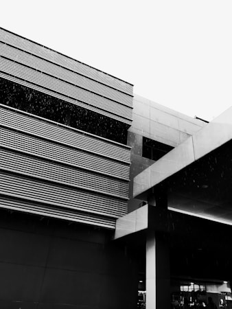 A modern building with horizontal slats and a minimalist design. The image is in black and white, highlighting the architectural lines and angles. Rain is falling, creating a textured effect.