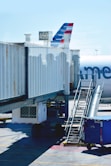 A commercial airplane is stationary at the gate of an airport. The aircraft's tail, with distinctive red, white, and blue stripes, is partially visible. Boarding stairs are positioned beside a large covered jet bridge connected to gate A50. The shadows of several people are visible on the jet bridge, and there is a blue container or baggage cart on the ground nearby.