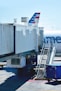 A commercial airplane is stationary at the gate of an airport. The aircraft's tail, with distinctive red, white, and blue stripes, is partially visible. Boarding stairs are positioned beside a large covered jet bridge connected to gate A50. The shadows of several people are visible on the jet bridge, and there is a blue container or baggage cart on the ground nearby.