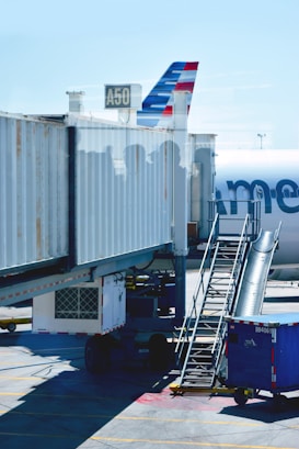 A commercial airplane is stationary at the gate of an airport. The aircraft's tail, with distinctive red, white, and blue stripes, is partially visible. Boarding stairs are positioned beside a large covered jet bridge connected to gate A50. The shadows of several people are visible on the jet bridge, and there is a blue container or baggage cart on the ground nearby.