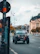 A Jeep vehicle drives down a city street lined with ornate historical buildings and street lamps. In the background, people are walking on the sidewalk, and trees with golden leaves add a splash of color to the urban landscape. The sky is cloudy, giving a diffused lighting effect.