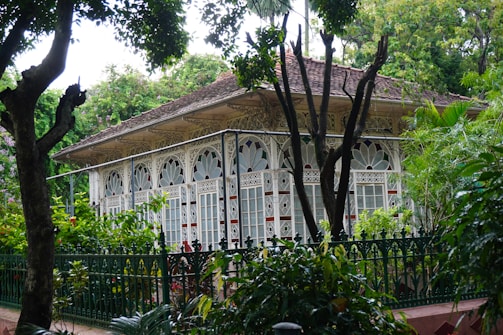 A beautifully decorated glass house with ornate detailing on the structure, surrounded by lush green foliage and trees. The building features intricate patterns and white framework, with large glass windows. The environment looks serene and natural.