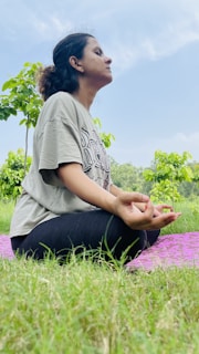 A person is seated cross-legged on a purple mat in a grassy area. The individual appears to be meditating, with closed eyes and hands resting on the knees in a mudra position. Lush green trees and a blue sky with some clouds are visible in the background.