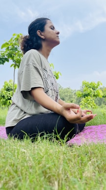 A person is seated cross-legged on a purple mat in a grassy area. The individual appears to be meditating, with closed eyes and hands resting on the knees in a mudra position. Lush green trees and a blue sky with some clouds are visible in the background.