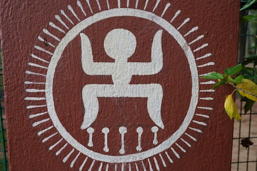 A tribal or traditional design painted on a brown surface features a white stylized human figure with arms raised and legs bent inside a circle. The circle is adorned with radiating white lines resembling rays or spikes.