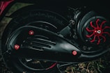 A close-up view of a motorcycle's mechanical components, including a section of the wheel, tire, engine, and exhaust cover. The engine has black metal parts with red accents and bolts giving it a rugged and industrial look. The background is slightly blurred, with some grass visible at the bottom of the image.