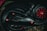 A close-up view of a motorcycle's mechanical components, including a section of the wheel, tire, engine, and exhaust cover. The engine has black metal parts with red accents and bolts giving it a rugged and industrial look. The background is slightly blurred, with some grass visible at the bottom of the image.