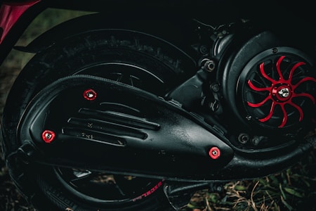 A close-up view of a motorcycle's mechanical components, including a section of the wheel, tire, engine, and exhaust cover. The engine has black metal parts with red accents and bolts giving it a rugged and industrial look. The background is slightly blurred, with some grass visible at the bottom of the image.