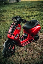 A vibrant red scooter is parked on a patch of grass. The scooter has a sleek design with black accents on the handlebars and footrests. The seat is black and appears cushioned for comfort. The environment around the scooter includes a grassy area with hints of sunlight filtering through, giving the scene a natural and serene backdrop.