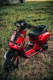 A vibrant red scooter is parked on a patch of grass. The scooter has a sleek design with black accents on the handlebars and footrests. The seat is black and appears cushioned for comfort. The environment around the scooter includes a grassy area with hints of sunlight filtering through, giving the scene a natural and serene backdrop.