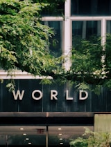 A building facade with large windows partially obscured by lush green foliage. The word 'WORLD' is prominently displayed on the building's exterior, possibly indicating the name or purpose of the building. The bottom section of the image shows a doorway with an 'EXIT' sign.