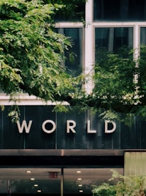 A building facade with large windows partially obscured by lush green foliage. The word 'WORLD' is prominently displayed on the building's exterior, possibly indicating the name or purpose of the building. The bottom section of the image shows a doorway with an 'EXIT' sign.