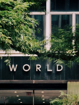 A building facade with large windows partially obscured by lush green foliage. The word 'WORLD' is prominently displayed on the building's exterior, possibly indicating the name or purpose of the building. The bottom section of the image shows a doorway with an 'EXIT' sign.