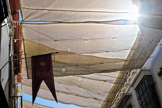 A street view looking upwards at a canopy made of beige and translucent fabric, stretched to provide shade. Sunlight peeks through on the right, creating a bright glare. A decorative maroon banner with text hangs on the left side. Buildings and metal gratings are partially visible on either side.