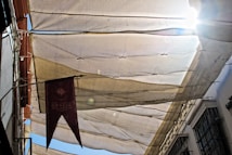 A street view looking upwards at a canopy made of beige and translucent fabric, stretched to provide shade. Sunlight peeks through on the right, creating a bright glare. A decorative maroon banner with text hangs on the left side. Buildings and metal gratings are partially visible on either side.