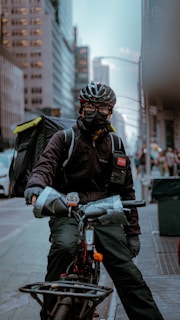 A bicyclist, wearing a helmet and face mask, is prominently positioned in an urban setting. They carry a large insulated backpack, indicating they might be a delivery worker. Tall buildings line the street, with soft lighting suggesting either early morning or late afternoon.