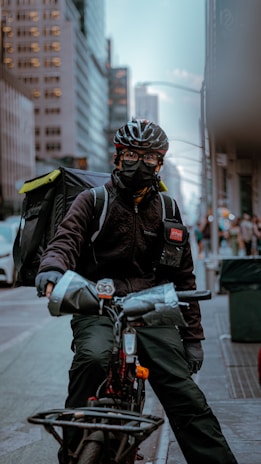 A bicyclist, wearing a helmet and face mask, is prominently positioned in an urban setting. They carry a large insulated backpack, indicating they might be a delivery worker. Tall buildings line the street, with soft lighting suggesting either early morning or late afternoon.