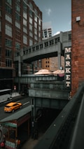 A street scene in an urban area with tall brick buildings and a connecting overhead walkway. A sign with the word 'MARKET' is prominently displayed on a building. Below, a street with a yellow taxi cab and a bus is visible, and a few pedestrians walk along the sidewalk. The atmosphere combines modern architecture with an industrial touch.