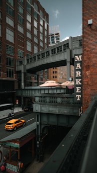 A street scene in an urban area with tall brick buildings and a connecting overhead walkway. A sign with the word 'MARKET' is prominently displayed on a building. Below, a street with a yellow taxi cab and a bus is visible, and a few pedestrians walk along the sidewalk. The atmosphere combines modern architecture with an industrial touch.