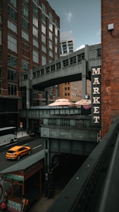 A street scene in an urban area with tall brick buildings and a connecting overhead walkway. A sign with the word 'MARKET' is prominently displayed on a building. Below, a street with a yellow taxi cab and a bus is visible, and a few pedestrians walk along the sidewalk. The atmosphere combines modern architecture with an industrial touch.