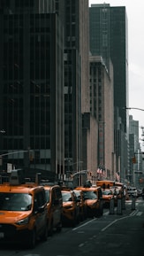 A row of yellow taxis lines a city street flanked by tall, dark skyscrapers. American flags are visible, and a sign for Radio City adds to the urban atmosphere.