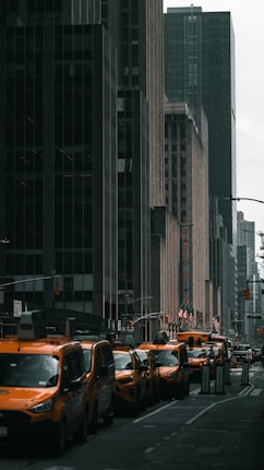 A row of yellow taxis lines a city street flanked by tall, dark skyscrapers. American flags are visible, and a sign for Radio City adds to the urban atmosphere.
