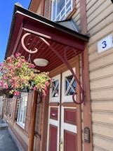 A classic wooden building with tan horizontal siding and red accents, featuring a prominent entrance door with geometric glass panels. A decorative metal bracket holds a hanging basket overflowing with vibrant pink flowers. A small blue and white house number plate with the numeral '3' is mounted near the door.