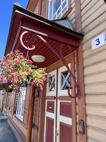 A classic wooden building with tan horizontal siding and red accents, featuring a prominent entrance door with geometric glass panels. A decorative metal bracket holds a hanging basket overflowing with vibrant pink flowers. A small blue and white house number plate with the numeral '3' is mounted near the door.