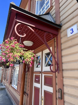 A classic wooden building with tan horizontal siding and red accents, featuring a prominent entrance door with geometric glass panels. A decorative metal bracket holds a hanging basket overflowing with vibrant pink flowers. A small blue and white house number plate with the numeral '3' is mounted near the door.
