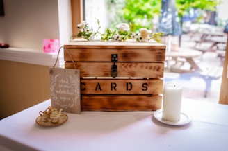 A wooden box labeled 'CARDS' is adorned with white flowers and greenery on top. Placed on a white tablecloth, the setup includes a tall white candle on a saucer to the right. To the left, a small sign reads 'And so our adventure begins,' alongside a miniature tea set.