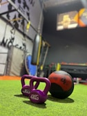 Two purple kettlebells labeled 4 KG each and a black and red medicine ball are positioned on a green mat in a gym setting. In the background, there is a punching bag and various gym equipment. The environment is slightly out of focus, with a mural on the wall and exercise tools scattered around.