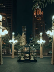 A small glass greenhouse is situated in the foreground surrounded by urban architecture and tall, ornate street lamps. The scene is set at night with the city skyline visible in the background, highlighted by a tall, illuminated gothic-style building. Potted plants line the interior of the greenhouse, and the pavement is clean and expansive.