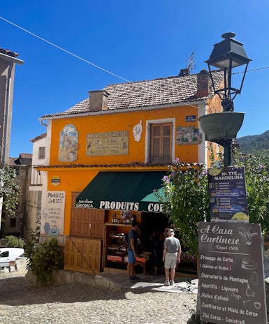 A charming, small store with a bright orange facade displaying various signs. The store has a green awning labeled 'Produits Corses' and sells local products. Two people are standing near the entrance, likely customers. The setting is sunlit with a clear blue sky, surrounded by stone buildings and lush greenery.