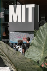 A large sign with bold letters spelling 'MIT' stands above a detailed campus map. Two individuals with backpacks are examining the map. Large green leaves frame the scene, adding a natural element to the modern setting.