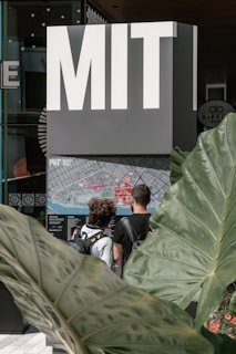A large sign with bold letters spelling 'MIT' stands above a detailed campus map. Two individuals with backpacks are examining the map. Large green leaves frame the scene, adding a natural element to the modern setting.