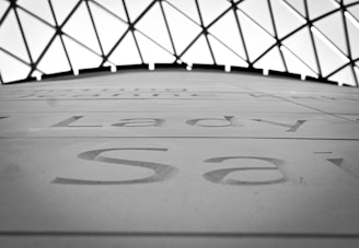 A close-up view of a surface with engraved letters, viewed from a low angle. The background features a geometric pattern of interconnected triangles resembling a modern architectural ceiling.