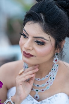 A woman with styled dark hair and makeup, wearing intricate gemstone jewelry. She rests her hand gently on her chin, exuding elegance and grace. Her makeup includes bold eye shadow, defined eyebrows, and a soft lip color. The background is softly blurred, emphasizing her as the focal point.