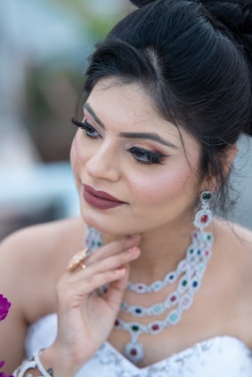 A woman with styled dark hair and makeup, wearing intricate gemstone jewelry. She rests her hand gently on her chin, exuding elegance and grace. Her makeup includes bold eye shadow, defined eyebrows, and a soft lip color. The background is softly blurred, emphasizing her as the focal point.
