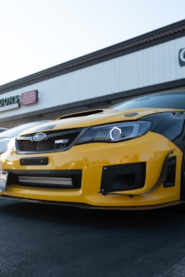 A close-up view of the front section of a yellow and black Subaru WRX parked in a commercial area. The car's hood features aggressive styling with a scoop, and the headlights have a modern, angular design. There are modest buildings in the background, with visible signs.