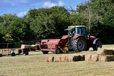 A red tractor is operating in a field, towing a hay baler that is processing the cut grass into rectangular bales. The bales are neatly arranged on the ground in the foreground. Tree-lined greenery creates a natural backdrop, and the sky is partly cloudy with blue patches visible.