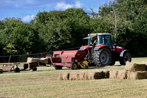 A red tractor is operating in a field, towing a hay baler that is processing the cut grass into rectangular bales. The bales are neatly arranged on the ground in the foreground. Tree-lined greenery creates a natural backdrop, and the sky is partly cloudy with blue patches visible.