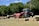 A red tractor is operating in a field, towing a hay baler that is processing the cut grass into rectangular bales. The bales are neatly arranged on the ground in the foreground. Tree-lined greenery creates a natural backdrop, and the sky is partly cloudy with blue patches visible.