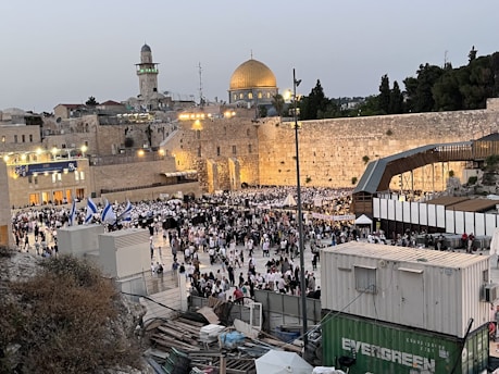 A large crowd of people gathered near the Western Wall in the Old City of Jerusalem. The Dome of the Rock is visible in the background, along with various architectural structures. People appear to be dressed in formal or traditional attire, and several Israeli flags are waving in the crowd.