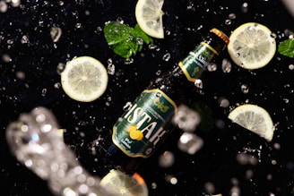 A beer bottle labeled with green and lemon graphics lies on a black surface surrounded by slices of lemon and mint leaves. Water droplets are splashing around, suggesting freshness and dynamism.